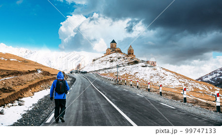 Stepantsminda, Georgia. Man Tourist Backpacker Traveler Photographer Walking To Holy Trinity Church - Tsminda Sameba. Concept Of Pilgrim Or Tourist Traveler. Bright Blue Cloudy Sky Above Church 99747133