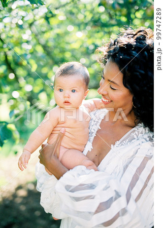 Smiling woman in white dress holding newborn baby in green garden 99747289