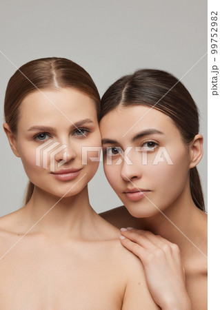 Closeup portrait of two adorable young girls with well-kept skin posing over grey background. Models with bare shoulders. Concept of beauty, spa, skin care and health Closeup portrait of two adorable young girls with well-kept skin posing over grey background. Models with bare shoulders. Concept of beauty, spa, skin care and health 99752982