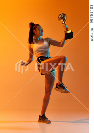 Success, win. Young sportive girl, professional athlete posing with trophy over orange studio background in neon light. Concept of sport. health and strength Success, win. Young sportive girl, professional athlete posing with trophy over orange studio background in neon light. Concept of sport. health and strength 99761483