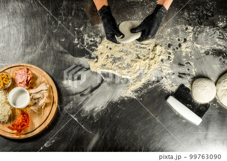 Cooking pizza on a steel table; dough, flour, pizza topping, sauce and chef`s hands in black gloves on the table Cooking pizza on a steel table; dough, flour, pizza topping, sauce and chef`s hands in black gloves on the table 99763090