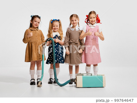 Little beautiful girls, kids in retro dresses playing with vacuum cleaner against grey studio background. Concept of childhood Little beautiful girls, kids in retro dresses playing with vacuum cleaner against grey studio background. Concept of childhood 99764647
