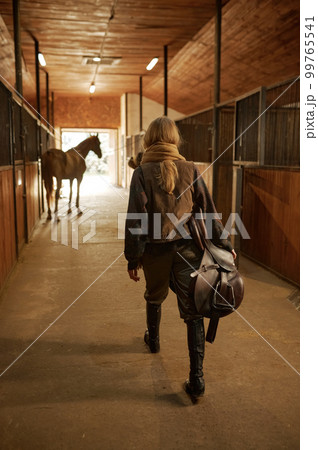 View from back on walking horsewoman carrying saddle in hand 99765541