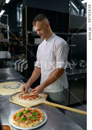 Pizzaiolo adding ham on pizza base with cheese, preparing italian dish 99765638