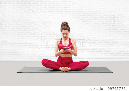 Attractive young woman in red sportswear sitting on a mat and choose music on her mobile in loft fitness studio. Attractive young woman in red sportswear sitting on a mat and choose music on her mobile in loft fitness studio. 99765775