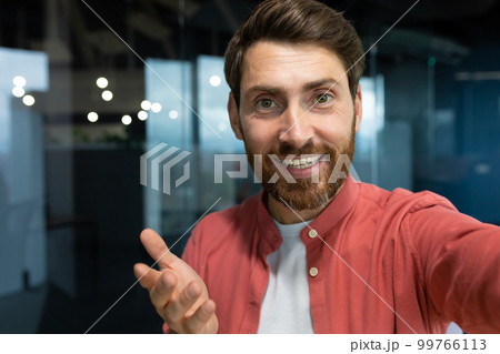 Close-up photo. Young businessman, freelancer, blogger in a red shirt in the office talking on a video call from the phone, smiling at the camera. 99766113