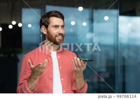 A young man in a red shirt sits in the office and talks on the phone through the loudspeaker, records the conversation on the recorder, voice message, looks away, smiles. A young man in a red shirt sits in the office and talks on the phone through the loudspeaker, records the conversation on the recorder, voice message, looks away, smiles. 99766114