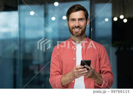 Portrait of a young man in a red shirt standing in the office, holding a phone in his hands, looking at the camera with a smile. 99766115