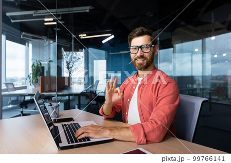 Portrait of a young male programmer, a freelancer who is reading in the office, sitting behind a computer, working on a laptop. He looks at the camera, smiles, shows with his hand ok. 99766141