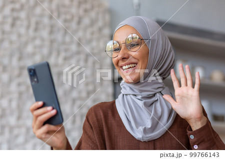 Young beautiful Muslim woman in hijab and glasses talking on video call from phone at home, waving at camera, greeting. Young beautiful Muslim woman in hijab and glasses talking on video call from phone at home, waving at camera, greeting. 99766143