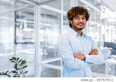 Young successful hispanic businessman in the middle of the office, portrait of happy and satisfied office worker, man in shirt and arms crossed smiling and looking at camera, businessman at work. Young successful hispanic businessman in the middle of the office, portrait of happy and satisfied office worker, man in shirt and arms crossed smiling and looking at camera, businessman at work. 99767651