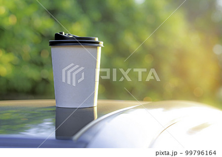 coffee takeaway in a paper cup on top of the car roof green tree background at sunrise in the morning,  selective focus, soft focus. 99796164