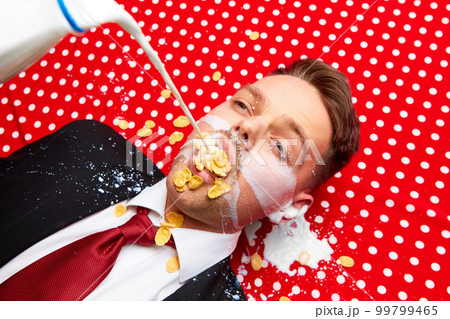 Young man lying on red vintage table cloth with corn flakes in mouth. Milk pouring inside mouth. Breakfast time. Food pop art photography. Young man lying on red vintage table cloth with corn flakes in mouth. Milk pouring inside mouth. Breakfast time. Food pop art photography. 99799465