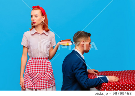 Young redhead woman serving breakfast with fried eggs and sausages to man against blue studio background. Food pop art photography. 99799468
