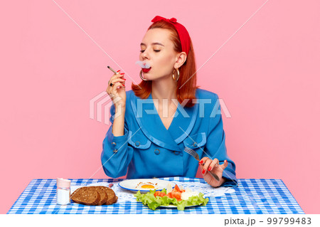 Stylish redhead woman sitting at table, smoking and having breakfast with fried eggs and salad against pink studio background. Food pop art photography. 99799483