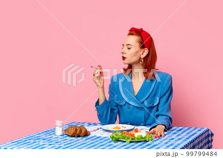 Stylish redhead woman sitting at table, smoking and having english breakfast with fried eggs against pink studio background. Food pop art photography. 99799484