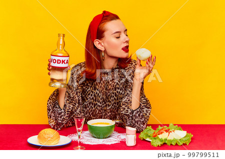 Food pop art photography. Beautiful redhead woman sitting at table against yellow background and having dinner, drinking vodka with onion. 99799511