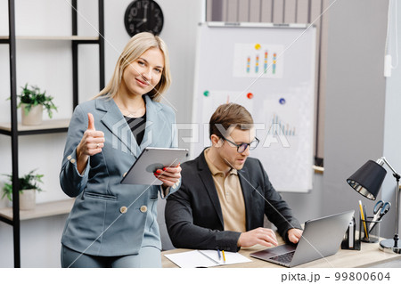 Cheerful young corporate lawyers in formal wear editing text of new contract using laptop sitting at table with laptop in modern office. Woman sits next to a colleague and holds a tablet in her hands 99800604