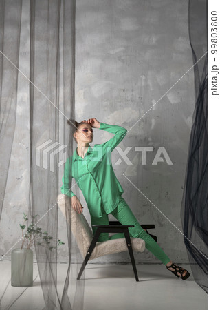 Stylish girl in green suit and black heels stands on gray wall background leaning on chair. Vertical frame Stylish girl in green suit and black heels stands on gray wall background leaning on chair. Vertical frame 99803800
