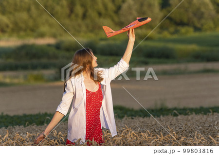 Happy young woman with toy airplane in hands in middle of wheat field, in red dress and white shirt Concept of cheap air travel Happy young woman with toy airplane in hands in middle of wheat field, in red dress and white shirt Concept of cheap air travel 99803816