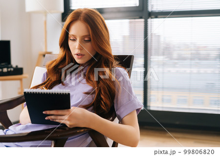 Close-up of focused young woman making notes in paper notebook sitting on comfy chair on background of window. Happy redhead female writing notes in diary or making to do list relaxing at home office Close-up of focused young woman making notes in paper notebook sitting on comfy chair on background of window. Happy redhead female writing notes in diary or making to do list relaxing at home office 99806820