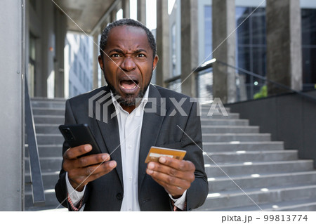 Worried shocked senior african american businessman in suit standing outside near office center. Holds a blocked credit card and phone. Bankruptcy, fraud. Screams angrily at the camera. 99813774