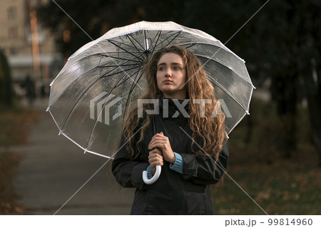 Brooding attractive woman with long hair walks through autumn park in rainy weather under transparent umbrella. Brooding attractive woman with long hair walks through autumn park in rainy weather under transparent umbrella. 99814960