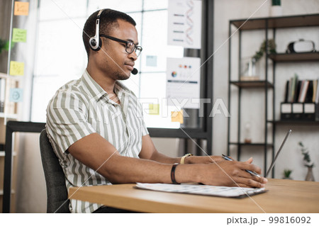 Busy african man using headset in striped shirt sitting at office and using wireless laptop. 99816092