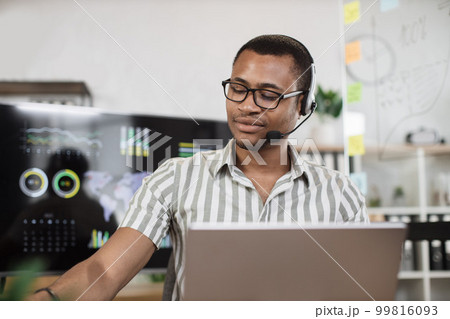 Confident african man in striped shirt using headset sitting at modern office and using laptop 99816093