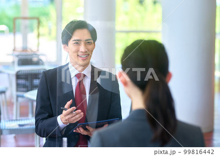 アクリル板のあるオフィスのロビーで面接するスーツ姿のビジネスマン 99816442