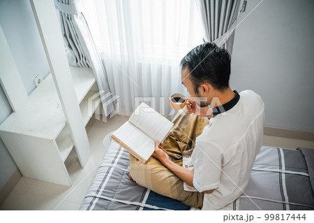 man reading book and drinking coffee while sitting on bed 99817444