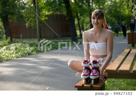 A young smiling girl sits on a bench in a city park with her rollerblades prepared. A young smiling girl sits on a bench in a city park with her rollerblades prepared. 99827102