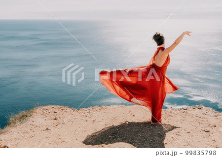 Woman in red dress on sea. Side view a Young beautiful sensual woman in a red long dress posing on a rock high above the sea on sunset. Girl on the nature on blue sky background. Fashion photo. Woman in red dress on sea. Side view a Young beautiful sensual woman in a red long dress posing on a rock high above the sea on sunset. Girl on the nature on blue sky background. Fashion photo. 99835798