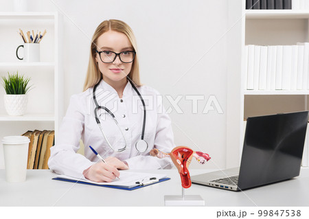 Young beautiful doctor woman working happy and smile in hospital, sitting on table. Model of female reproductive system on work desk. Young beautiful doctor woman working happy and smile in hospital, sitting on table. Model of female reproductive system on work desk. 99847538