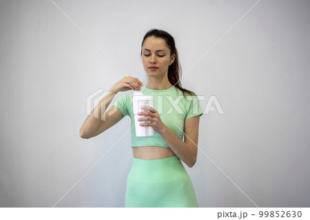 A young, slim woman in sportswear twisting the lid of a sports nutrition shaker against a gray background. Copy space. 99852630
