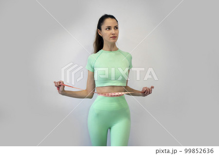 Young slender woman in sportswear measuring her waist with a tape measure against a gray background. Copy Space. The concept of diet and weight loss. 99852636