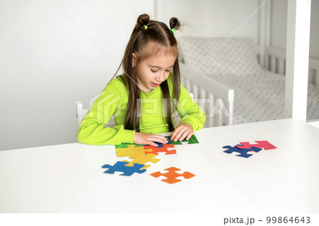 A girl in a bright light green T-shirt is sitting at a desk in the children's room, putting the colored puzzle pieces together. 99864643