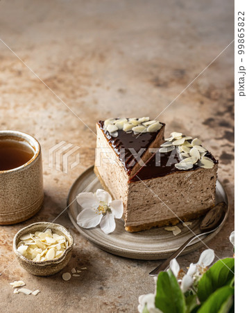 Two slices of chocolate cheesecake with almond petals, tea cup on brown textured background with copy space. Spring flowers in blur. Menu 99865822