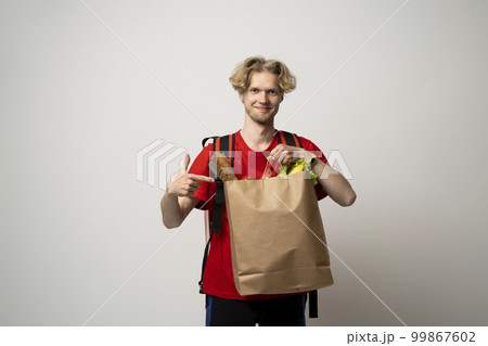 Food delivery service. Portrait of pleased delivery man in red uniform smiling while carrying paper bag with food products isolated over white background. 99867602