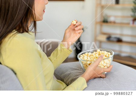 Young asian woman putting a bowl of popcorn on pillow and eating popcorn while sitting on the big comfortable Young asian woman putting a bowl of popcorn on pillow and eating popcorn while sitting on the big comfortable 99880327