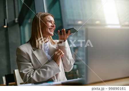 Happy businesswoman recording audio message on phone while while sitting in cozy cafe near windows 99890161