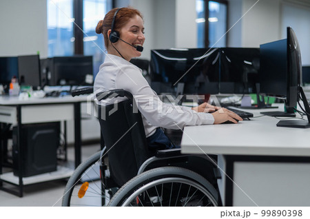 Red-haired caucasian woman in a wheelchair talking on a headset. Female call center worker smiling. 99890398