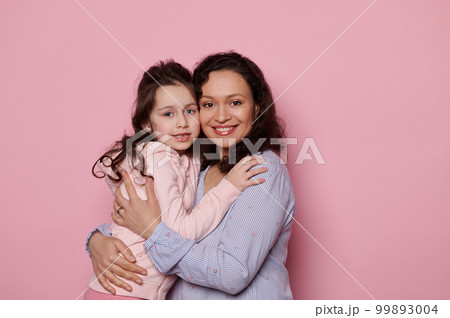 Loving mother hugging her little daughter, cutely smiling looking at camera, standing together on isolated pink background. Copy space. Happy family relationships. Mother's and Children's Day concept Loving mother hugging her little daughter, cutely smiling looking at camera, standing together on isolated pink background. Copy space. Happy family relationships. Mother's and Children's Day concept 99893004