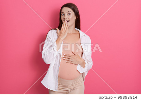 Young pregnant woman with expression of forgetting something isolated over rose background, keeping hand on belly, wearing leggins and casual shirt. 99893814
