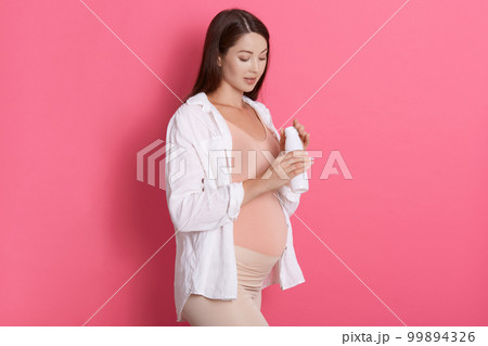 Pregnant woman holding bottle of milk in hands, expectant mother posing isolated over rosy wall, looking at her yogurt, wearing white shirt and leggins. 99894326