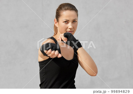 Photo of female boxer in defensive stance, looks fierce, dressed in black t shirt, bandages on hands, ready to fight with opponent, stands against over grey background. People and boxing concept Photo of female boxer in defensive stance, looks fierce, dressed in black t shirt, bandages on hands, ready to fight with opponent, stands against over grey background. People and boxing concept 99894829