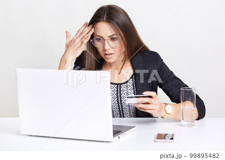 Surprised woman with dissatisfied expression, makes online transaction, works with laptop computer, makes shopping in web store, uses credit card, drinks glass of water, isolated on white wall Surprised woman with dissatisfied expression, makes online transaction, works with laptop computer, makes shopping in web store, uses credit card, drinks glass of water, isolated on white wall 99895482