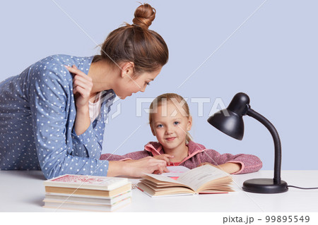 Smiling little charming girl sits at table and her mother help her to do homework task, try to learnn poem together, uses reading lamp for good vision, isolated over white studio background. Smiling little charming girl sits at table and her mother help her to do homework task, try to learnn poem together, uses reading lamp for good vision, isolated over white studio background. 99895549