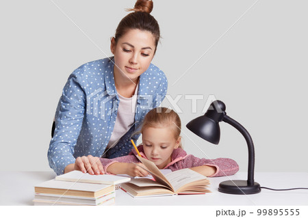 Photo of mother and daugher pose at desktop together, wrtite information in notebook, read many books, prepare for lessons at school, involved in studying, isolated over white studio background Photo of mother and daugher pose at desktop together, wrtite information in notebook, read many books, prepare for lessons at school, involved in studying, isolated over white studio background 99895555
