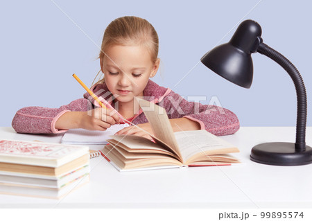 Close up portrait of little charming girl sits at white desk, does homework task, try to write composition or does sums, looks concentrated, uses reading lamp for good vision, isolated over blue wall. 99895574